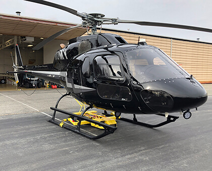 Black Eurocopter AS350 with red stripes parked on a tarmac in front of a white airplane hangar, being moved using a Chopper Spotter.