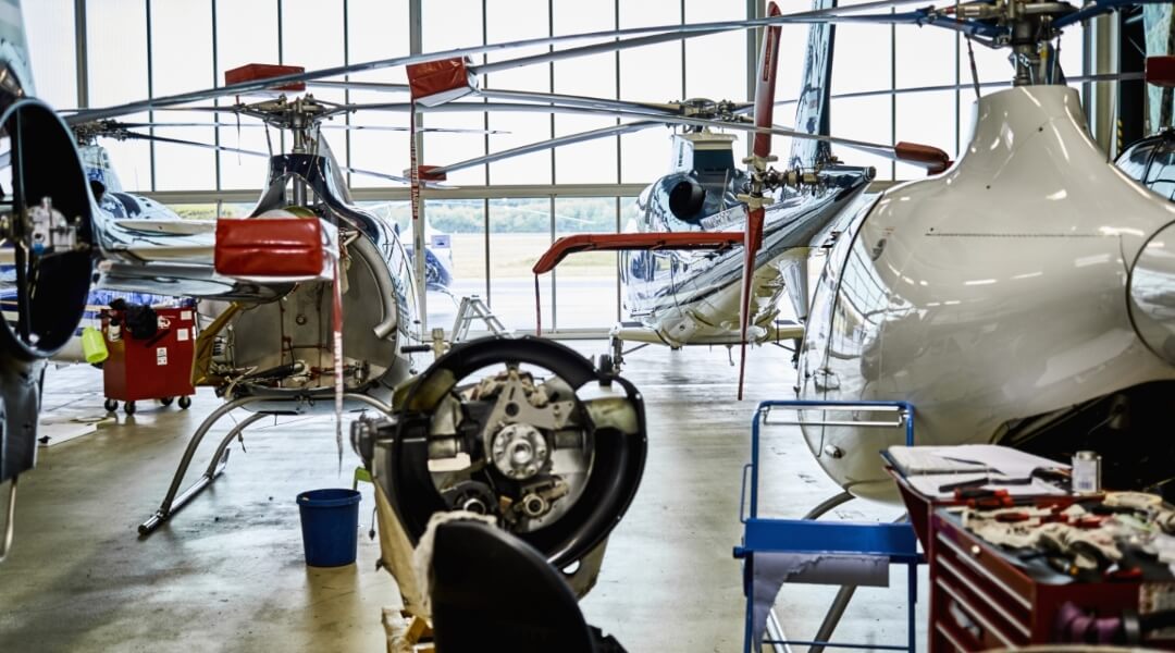A bustling maintenance hangar with a helicopter fleet in various stages of repair. Tools and equipment are scattered around, including a blue cart on the right and mechanical parts in the foreground. Large windows in the background allow natural light to fill the space, illuminating the scene.
