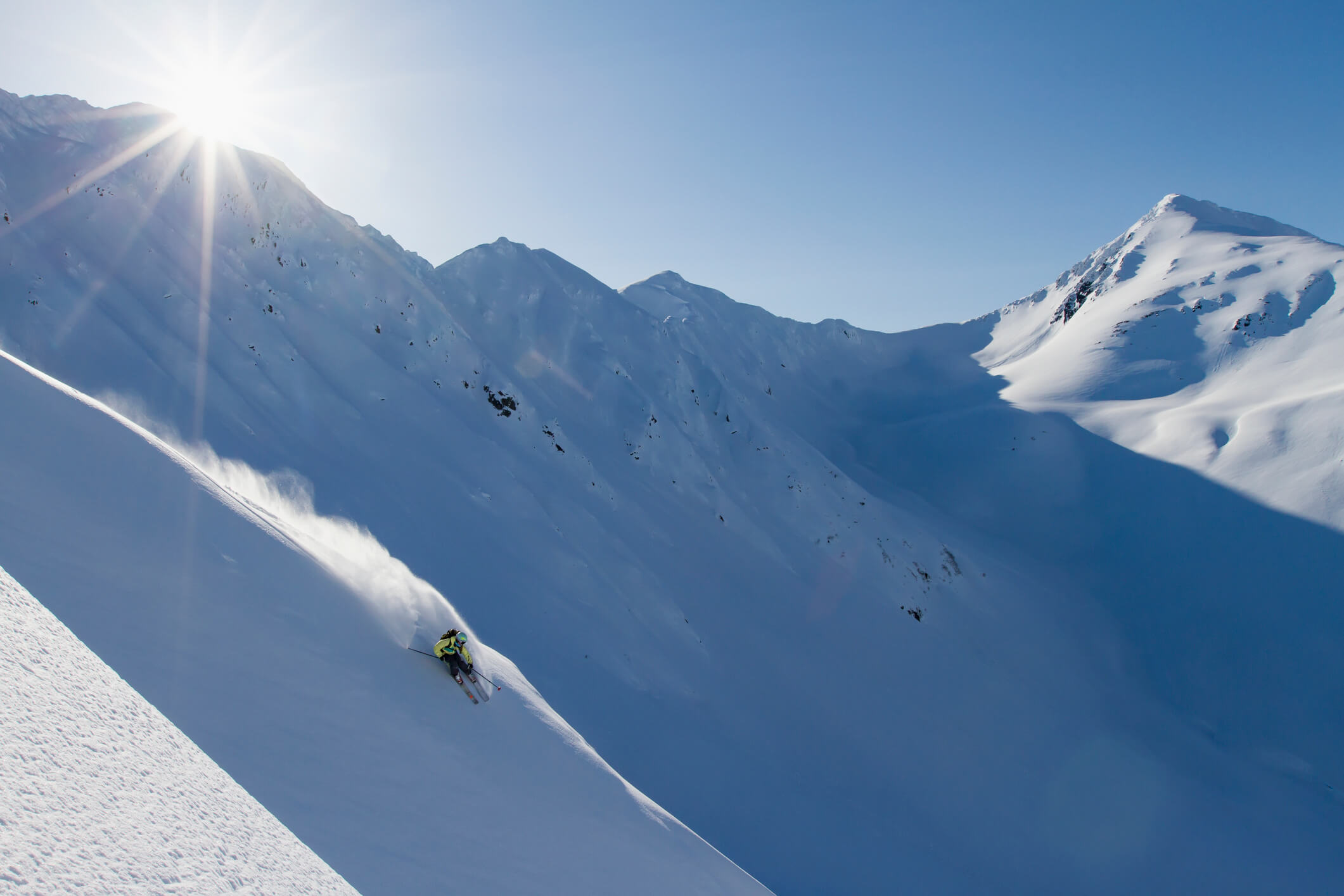 A skier in bright gear carves down a steep, snowy mountain slope—one of the best heli skiing locations—leaving a trail of powder as the sun shines above distant peaks, casting long shadows across the snow-covered landscape.
