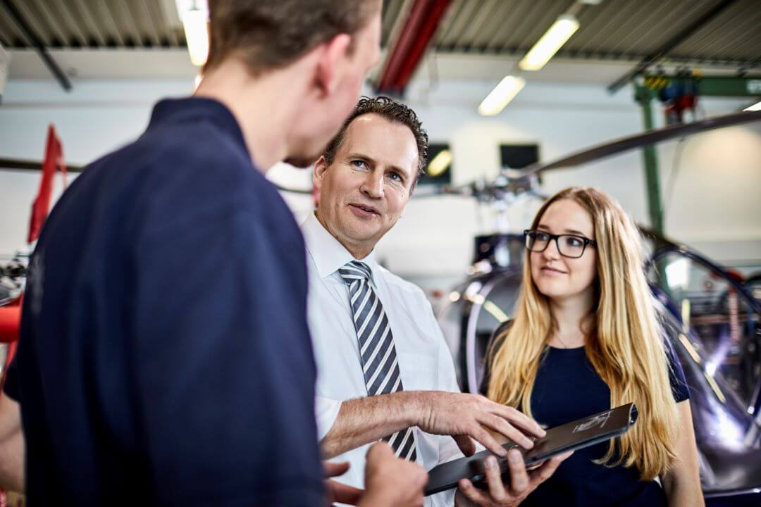 Three people are gathered in conversation, with two men and one woman. One man, dressed in a white shirt and striped tie, is holding a tablet and appears to be explaining something. The other man and the woman, both in casual attire, are listening attentively as they ask helicopter questions.
