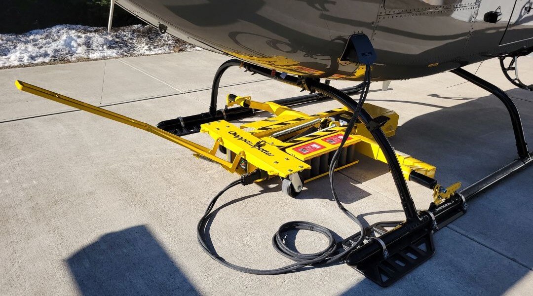 A yellow helicopter ground handling cart is positioned under a helicopter, ready to lift it for transport. The cart has wheels, control panels, and a long yellow handle, sitting on a concrete surface with some snow nearby.