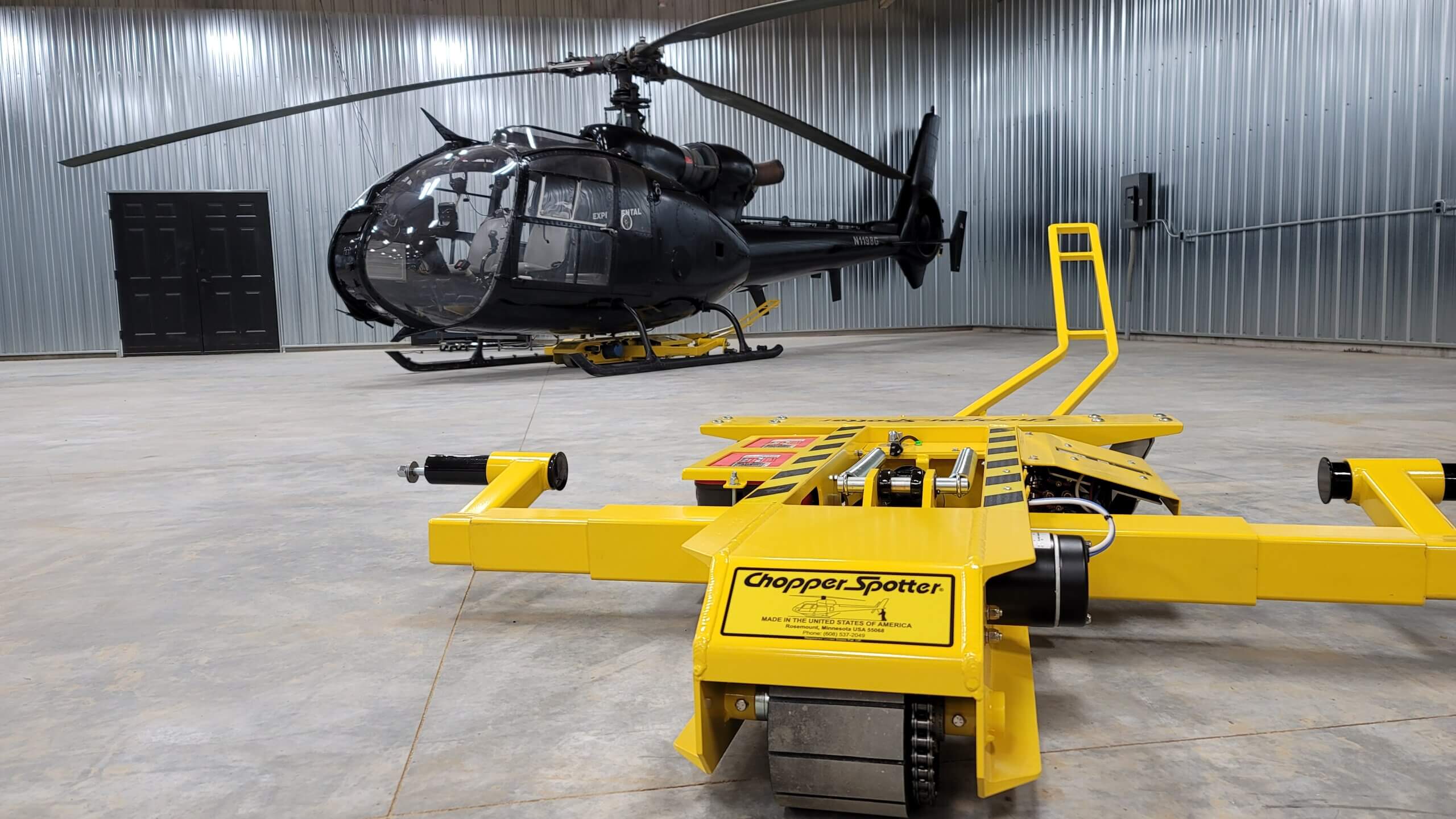 A black helicopter is parked inside a hangar with metal walls. In the foreground, a yellow "Chopper Spotter" device sits on the concrete floor, showcasing innovative personal helicopter moving solutions.