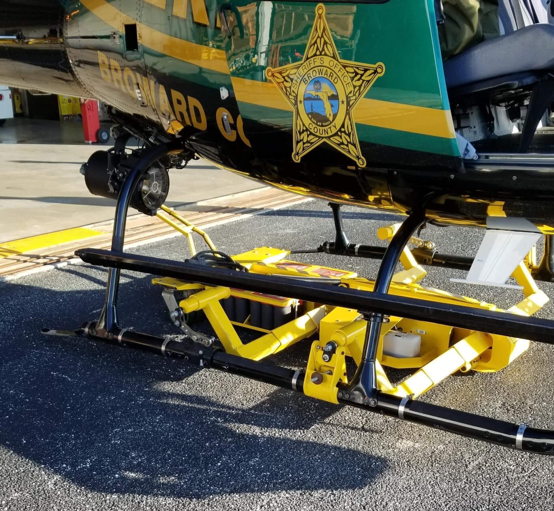 A Broward County Sheriff's Office helicopter is pictured on the ground, with its landing skids secured in a bright yellow police aviation ground support equipment cart designed for moving the aircraft.