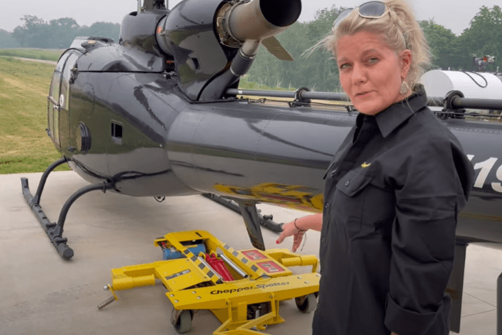A woman in a black shirt stands beside a helicopter on a concrete pad, pointing at a yellow 