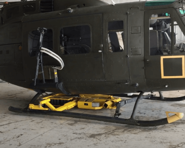 A green military helicopter parked inside a hangar has a yellow robotic device attached beneath its side, with a mounted gun and ammunition belt visible through the open door.