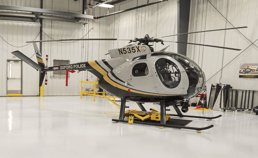 A small black, white, and gold helicopter marked “Oxford Police” and registration “N535X” is parked inside a well-lit, clean hangar with white walls, yellow safety barriers, and police aviation ground support equipment nearby.