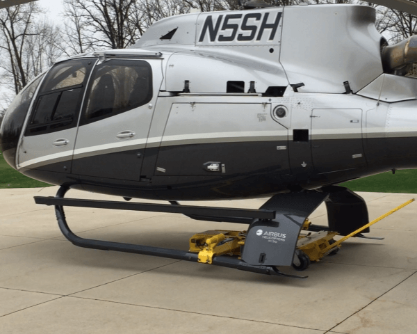 A gray and black Airbus helicopter with the tail number N5SH is parked on a paved surface, with yellow ground handling equipment positioned under its landing skids. Trees are visible in the background.