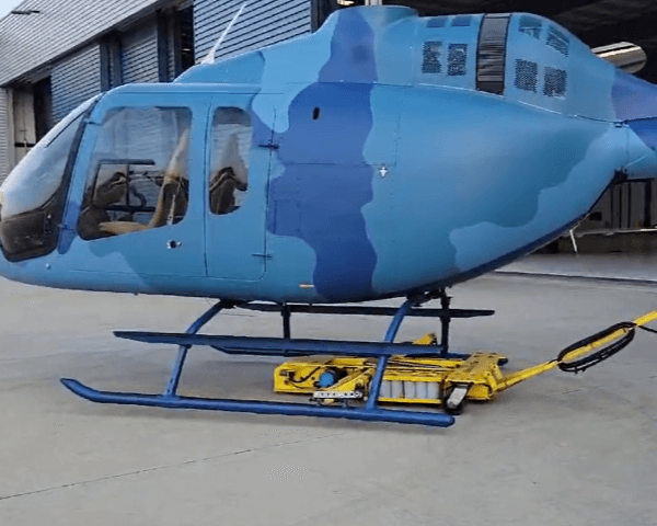 A blue helicopter with a camouflage pattern is parked on a concrete surface near a building, resting on a yellow aircraft tug. The helicopter has two windows and skids visible.