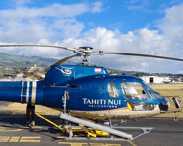 A blue Tahiti Nui helicopter is parked on an airstrip with grassy fields and mountains in the background. The sky is partly cloudy, and some airport equipment is visible near the helicopter.