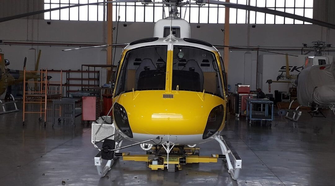 A yellow and white helicopter is parked inside a hangar, facing forward. The helicopter is undergoing maintenance, with panels open and equipment around it. The hangar floor is reflective and shelves are visible in the background.