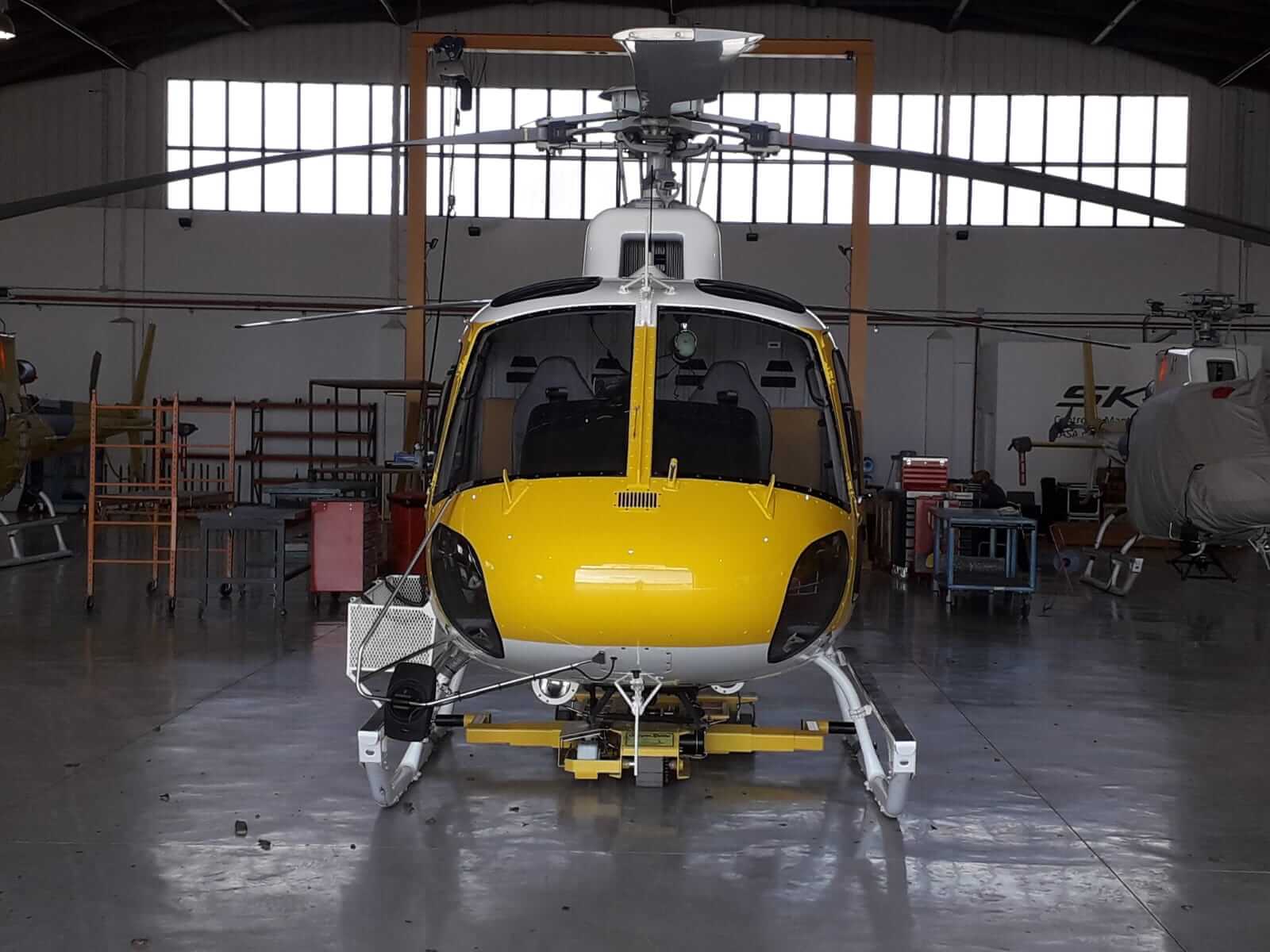 A yellow and white helicopter is parked inside a spacious hangar with polished concrete floors and various maintenance equipment around it. The helicopter is viewed from the front.