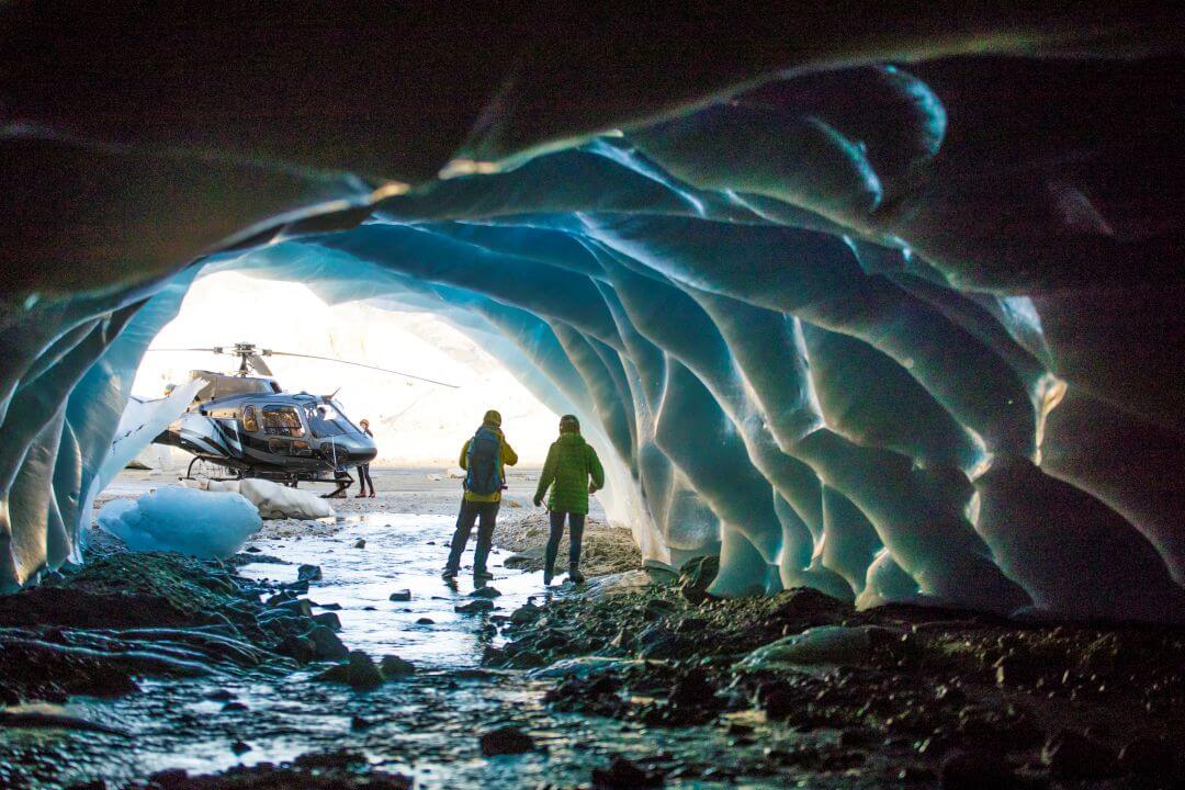 Two people stand inside an icy blue cave, looking out toward a helicopter parked on snowy ground outside. The cave walls are textured with ice formations, and sunlight filters in from the entrance.
