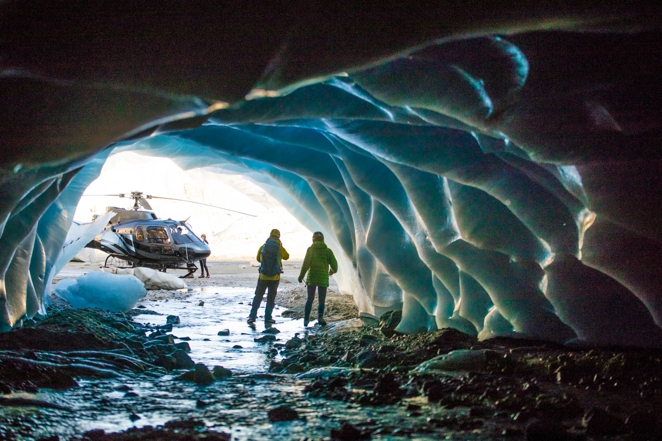 Two people stand inside a blue ice cave, looking out toward a helicopter parked on rocky ground outside the cave. Sunlight illuminates the icy cave walls and the scene outside.