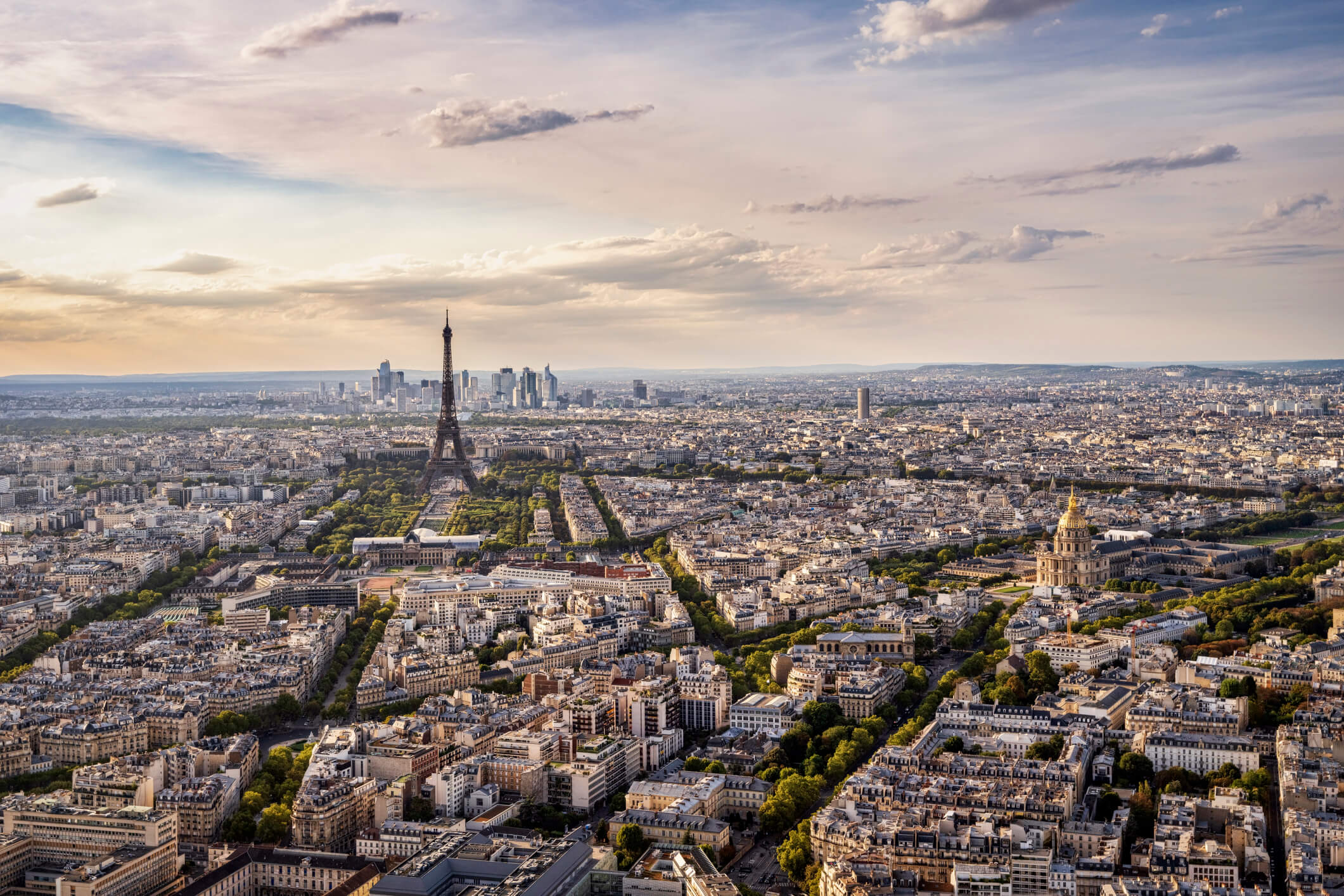 Aerial view of Paris at sunset, with the Eiffel Tower in the center, surrounded by city buildings, tree-lined streets, and a partly cloudy sky.