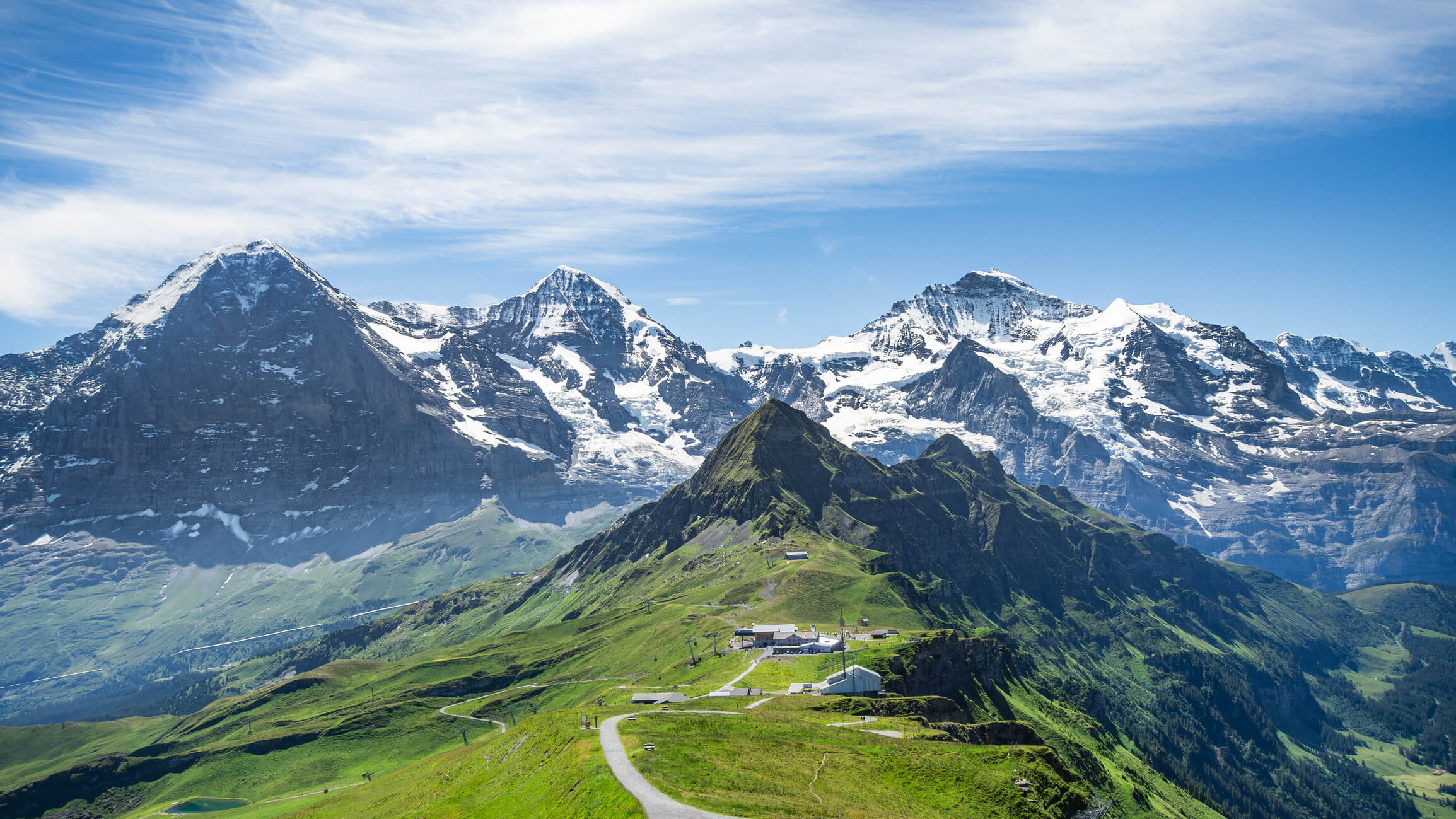 Picture shows the south facades of the most famous summits of the Bernese Alps, from left to right: the Eiger (3 967 m), the Mönch (4 110 m) and the Jungfrau (4 158 m) with the Jungfraujoch between the last two, the highest train station in Europe. The Tschuggen peak can be seen in front of them. The picture was taken from the Männlichen summit on the Panoramaweg hiking trail, also visible in the picture, linking the Männlichen (2 342 m) to the Kleine Scheidegg train station (2 061 m). It offers beautiful views of the surrounding summits and valleys, such as this one. The northern facades of the three peaks are classified as UNESCO World Heritage sites, as part of the inscription of the Jungfrau-Aletsch-Bietschhorn region in 2001.