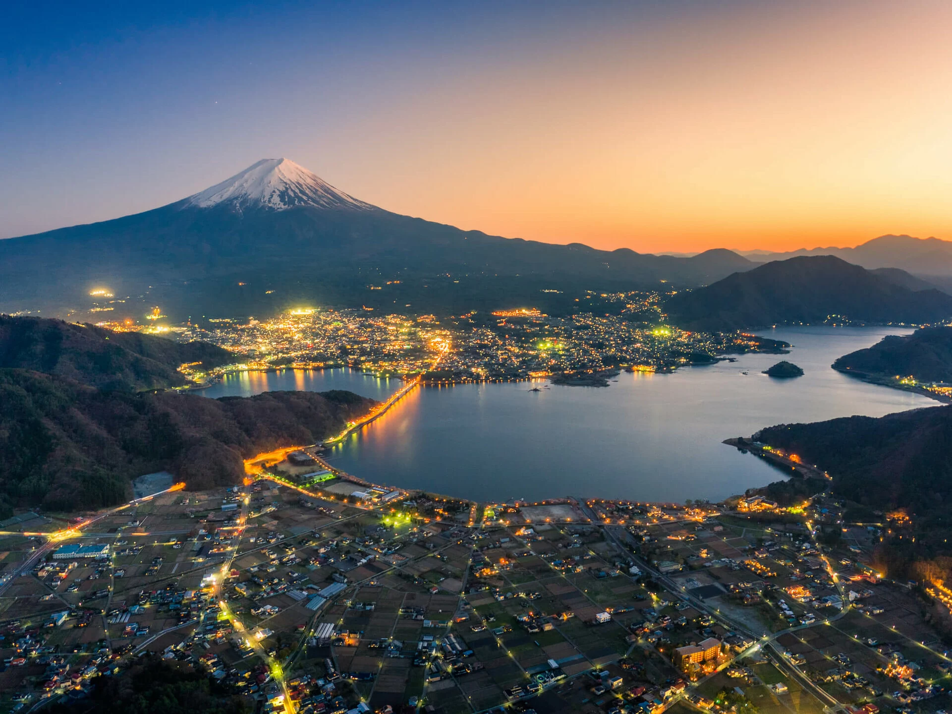 A scenic view of Mount Fuji at sunset with a town illuminated by lights along the shore of a large lake, surrounded by mountains and fields.