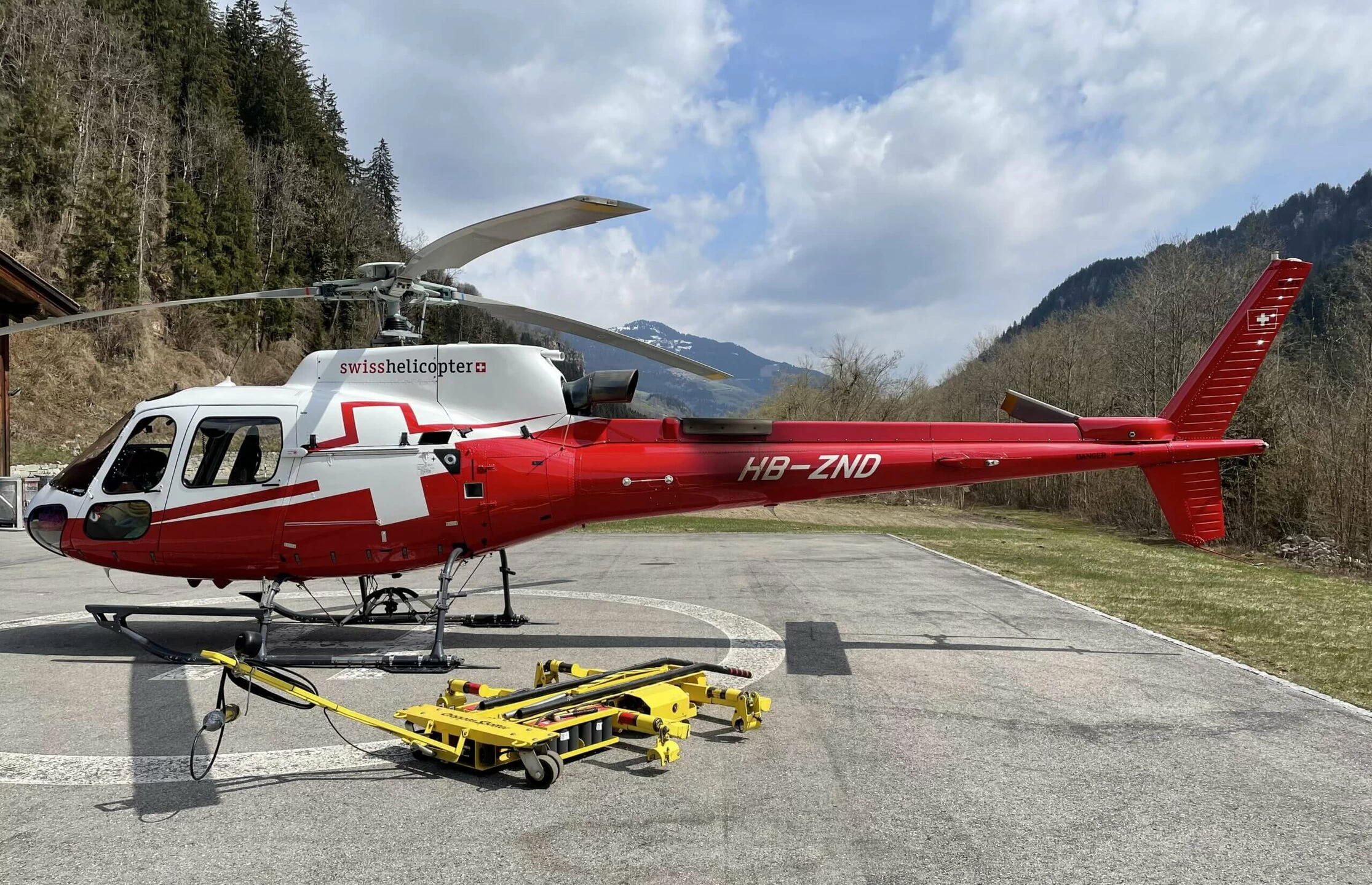 A red and white helicopter labeled "swisshelicopter" is parked on a helipad in a mountainous area, with a yellow ground-handling trolley positioned nearby and trees and hills in the background.