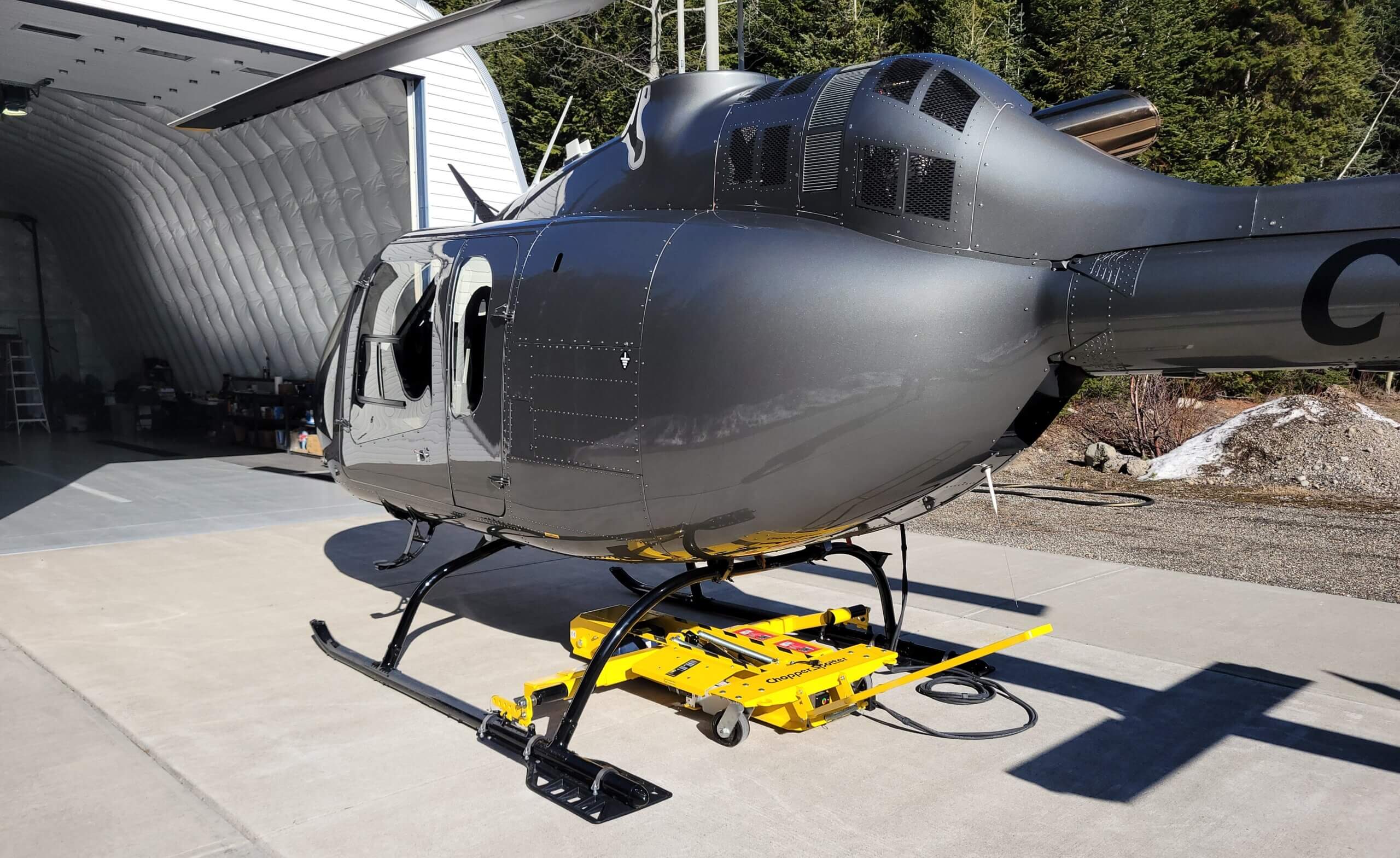 A sleek black helicopter is parked on a concrete surface outside a hangar, resting on a bright yellow ground handling wheel dolly, with trees visible in the background.