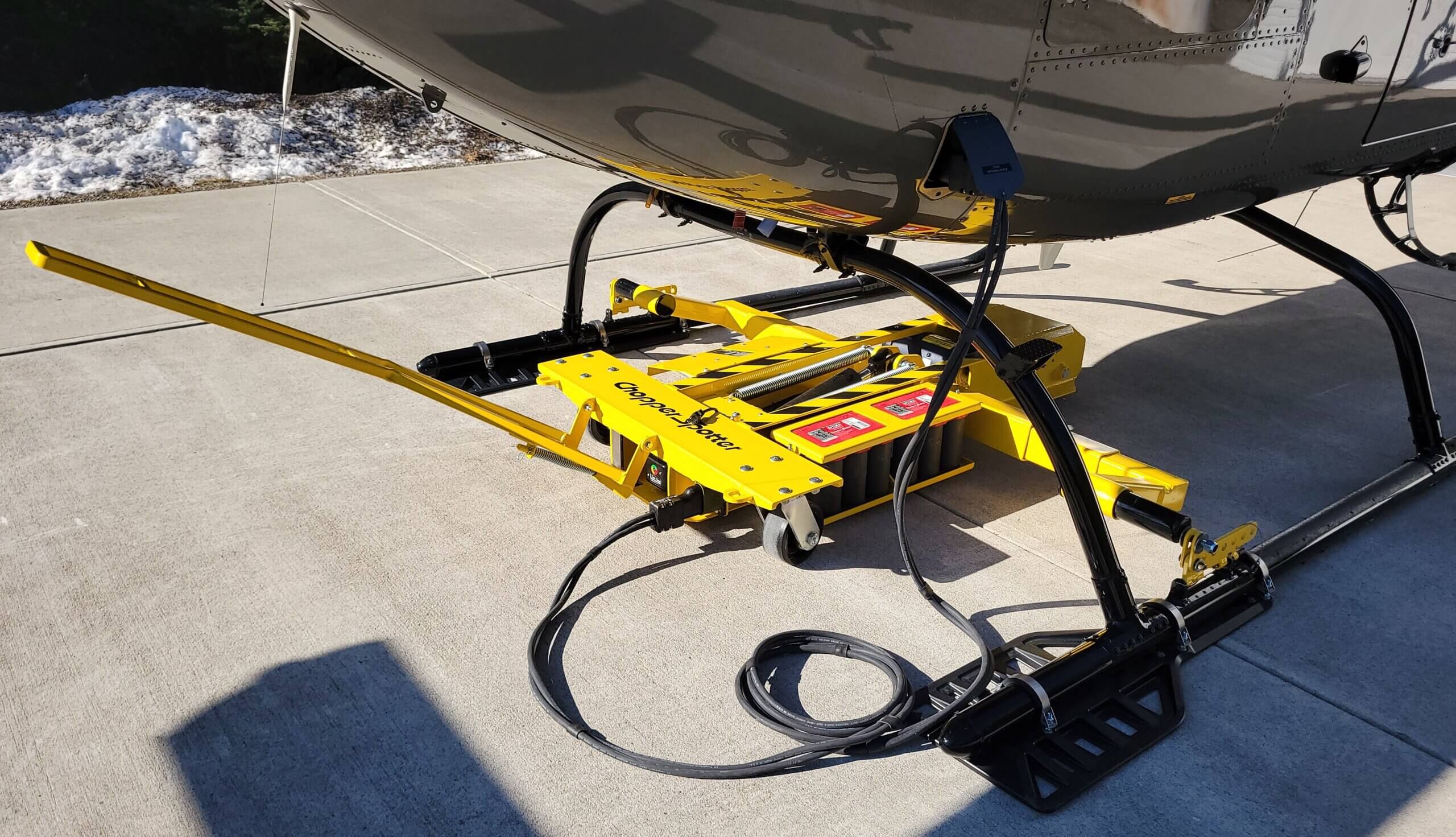 A yellow helicopter ground handling dolly is attached to the landing skids of a helicopter on a concrete surface, with a coiled cable and snow visible in the background.