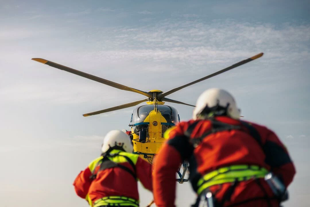 Two rescue workers in helmets and orange gear approach a yellow helicopter on a sunny day, preparing for a mission or emergency response. The helicopter’s rotors are visible against the sky.