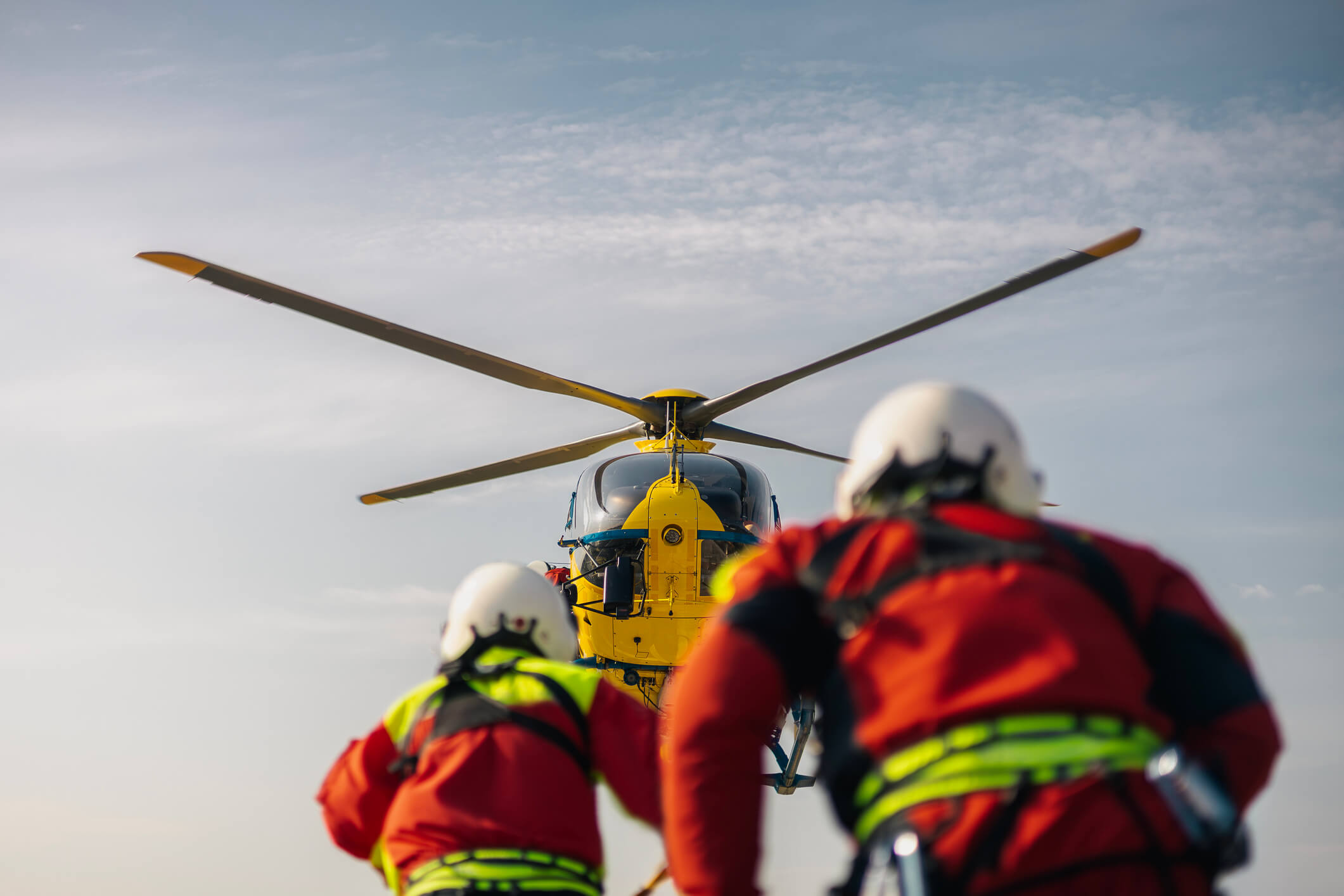 Two rescue workers in helmets and red suits approach a yellow helicopter from behind, set against a blue sky with wispy clouds.