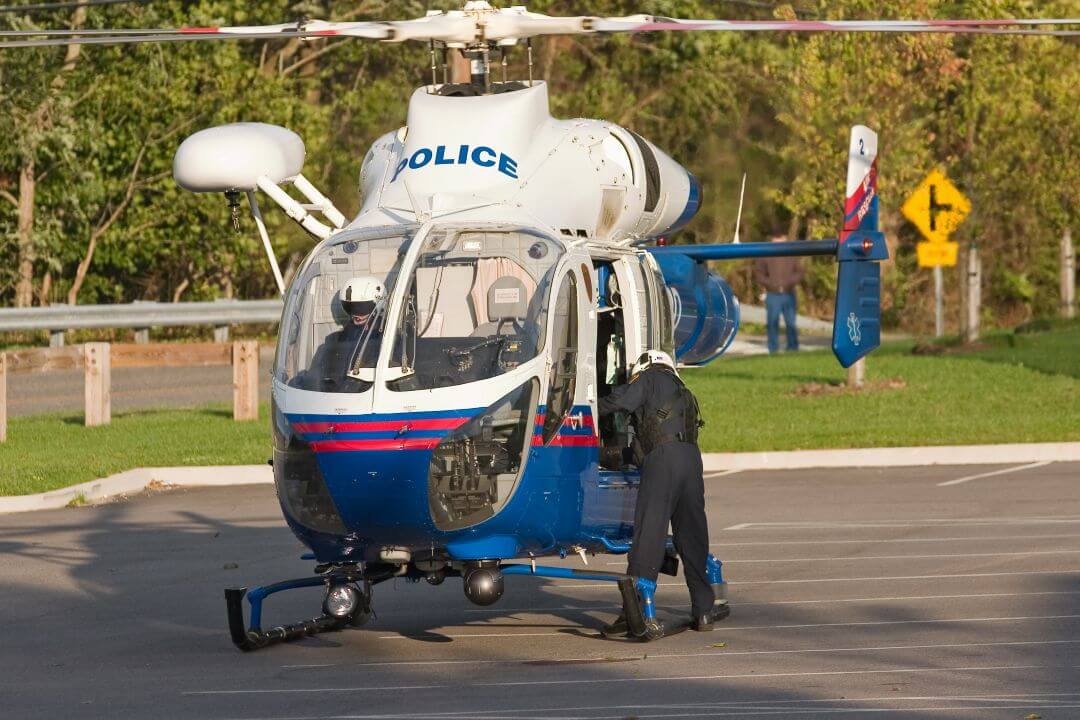 A police helicopter is parked on a paved lot. Two officers in uniform, one inside the cockpit and one standing outside near the open door, are preparing for or finishing a mission. Trees and a road sign are visible in the background.
