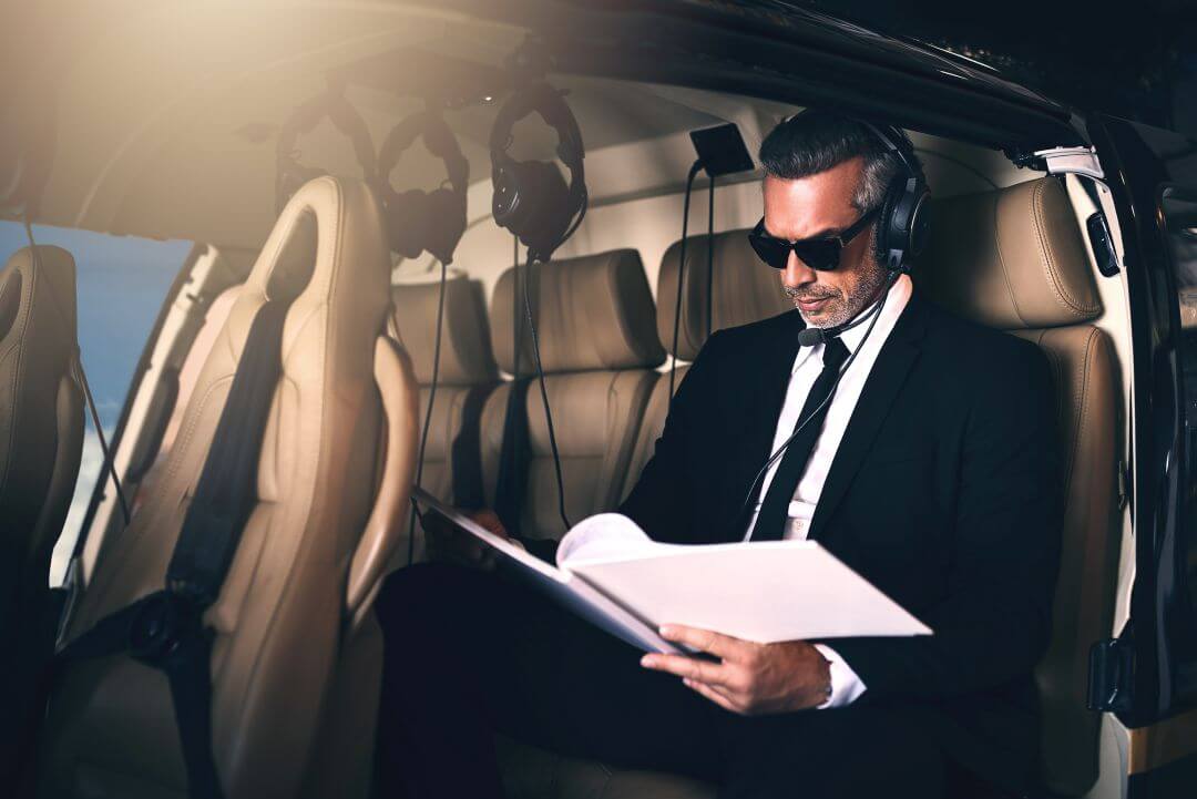 A man in a suit and sunglasses sits in a helicopter, wearing headphones and reading documents. Sunlight streams in, illuminating the luxurious beige leather seats.