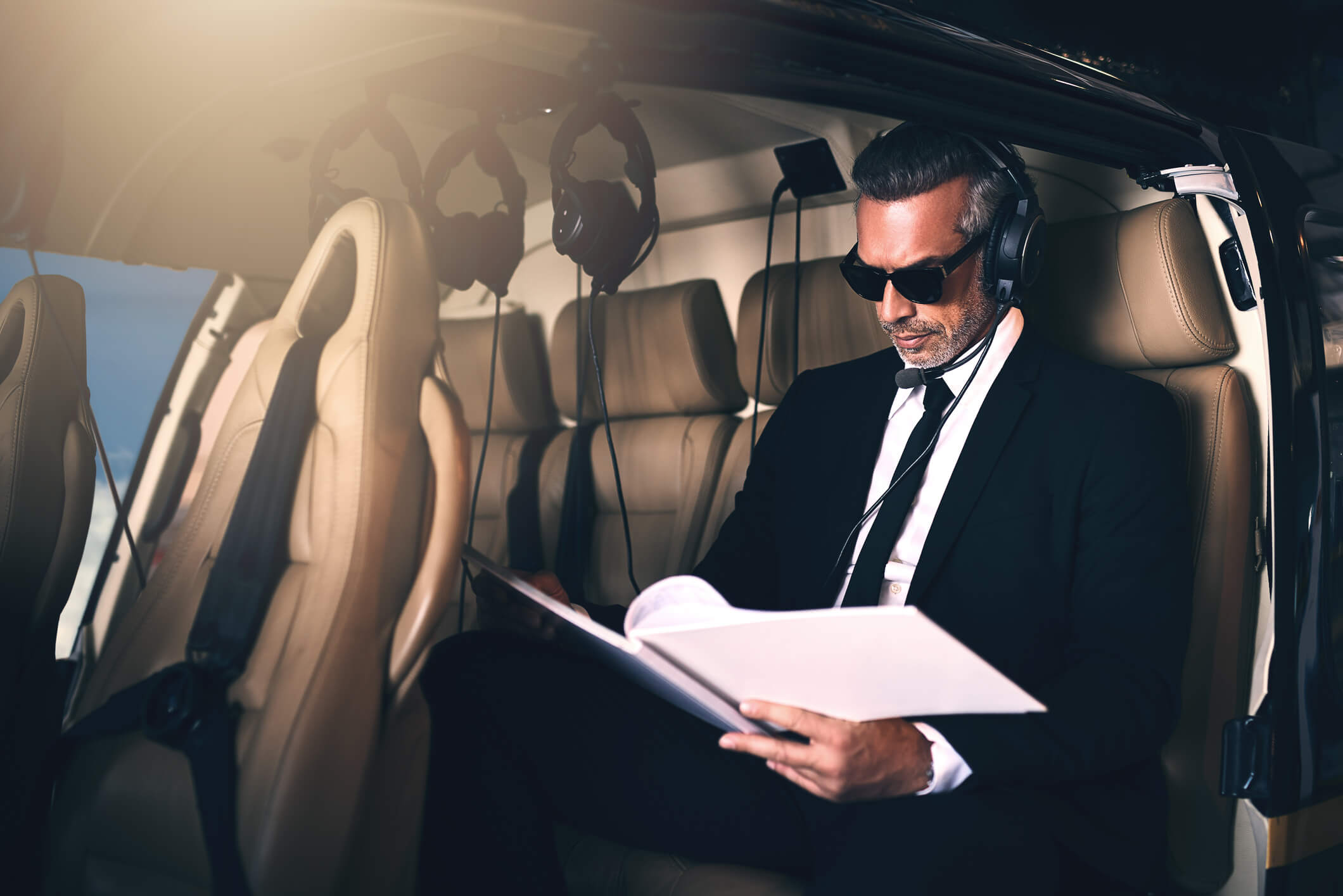 A man in a suit and sunglasses sits in a helicopter, wearing headphones and reading documents. Sunlight filters through the window, illuminating the beige leather seats and creating a focused, professional atmosphere.