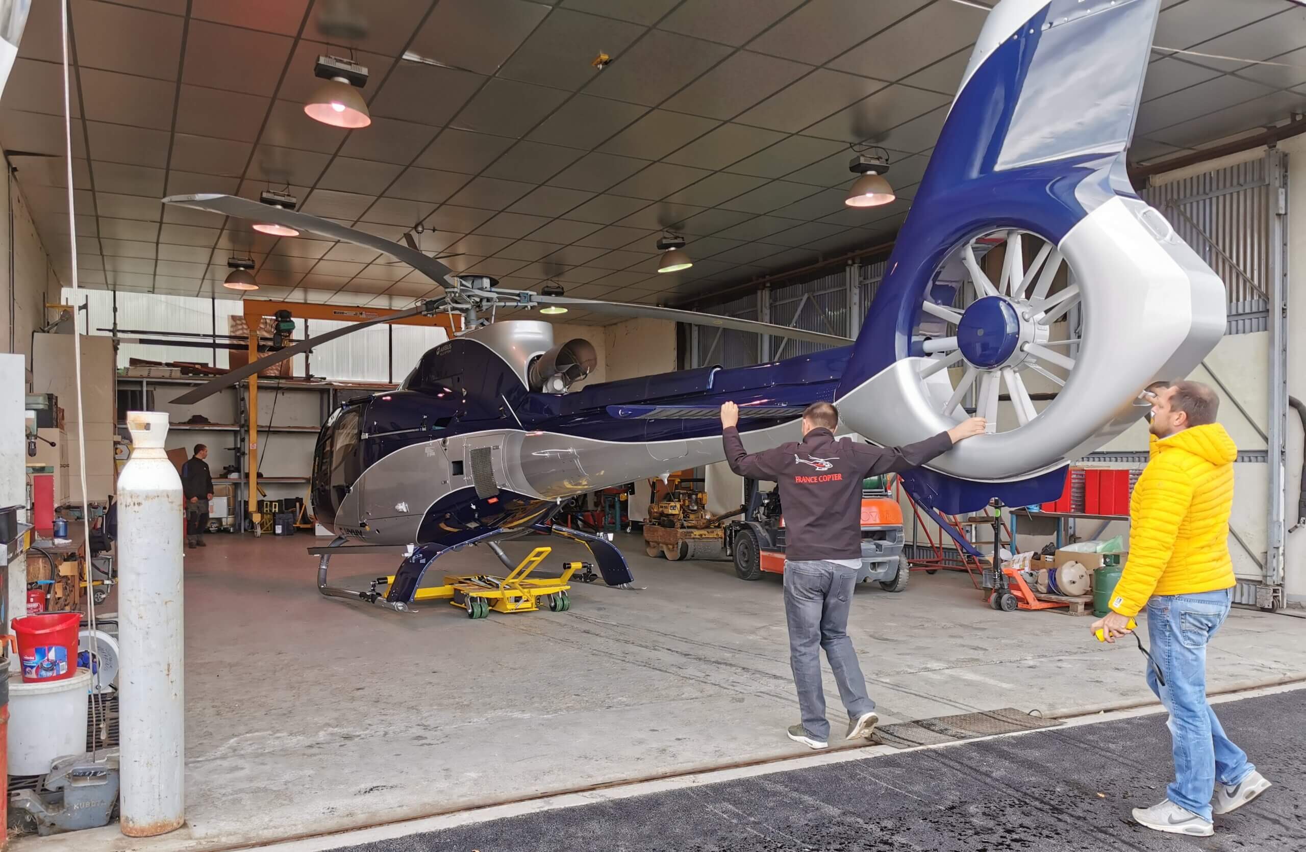 Two people stand near a blue and white helicopter inside a hangar. One person gestures toward the aircraft, while the other watches. The hangar contains tools and equipment.